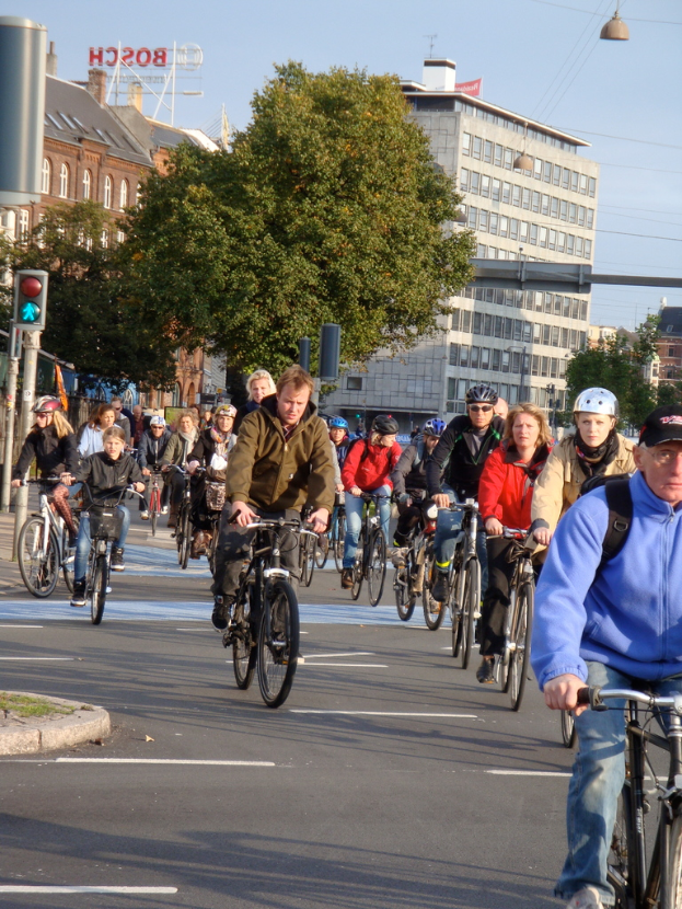 Menschen auf Fahrrädern auf einer Straße mit Pfählen, Bäumen, Gebäuden und dem Himmel im Hintergrund.