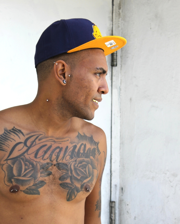 Eine Person mit einer Mütze mit einem Aufkleber mit einem Barcode und Bildern, mit sichtbaren Tattoos auf dem Körper, vor einer Tür und einer Wand stehend.