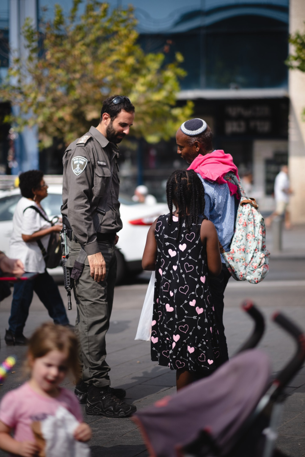 A police officer speaking to a group of people on a street, with a stroller in the foreground and vehicles, trees, and a building in the background.