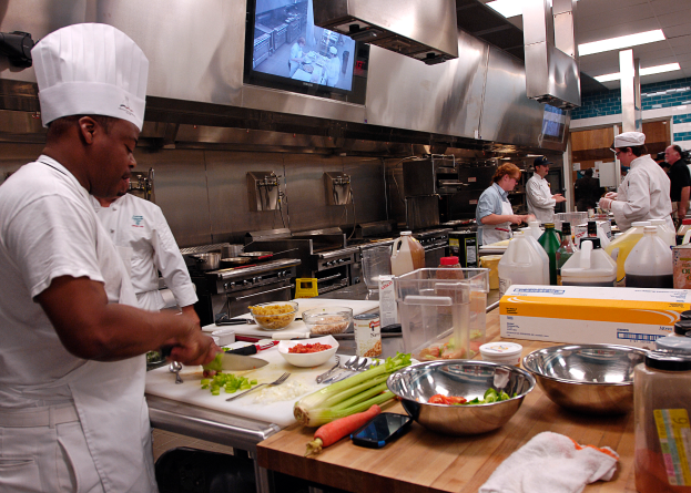 Eine Gruppe von Köchen in einer Küche, die Essen auf einem Schneidbrett zubereiten, mit einem Koch im Vordergrund in weißer Kleidung und Mütze, der ein Messer hält, und umgeben von verschiedenen Utensilien, Schüsseln, Gemüse, einem Handy, Flaschen und anderen Gegenständen, mit einem Fernseher und Deckenlampen im Hintergrund.
