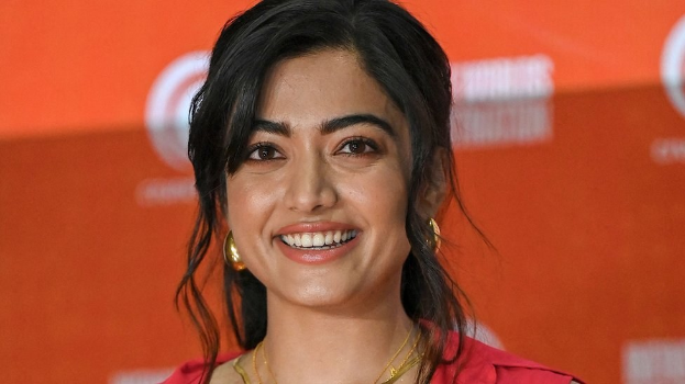 Eine Frau in einem roten Kleid mit einem eleganten Dutt, schwarzen High Heels und natürlichem Make-up lächelt in die Kamera und steht vor einem hellgelben Banner mit der Aufschrift "Sonam Kapoors beste Schönheits Looks vom Cannes Film Festival."
