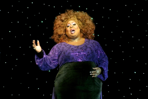 Eine Frau in einem lilafarbenen Kleid steht auf einer Bühne, beleuchtet von Hintergrundlichtern, als würde sie mitten in einer Vorstellung sein.