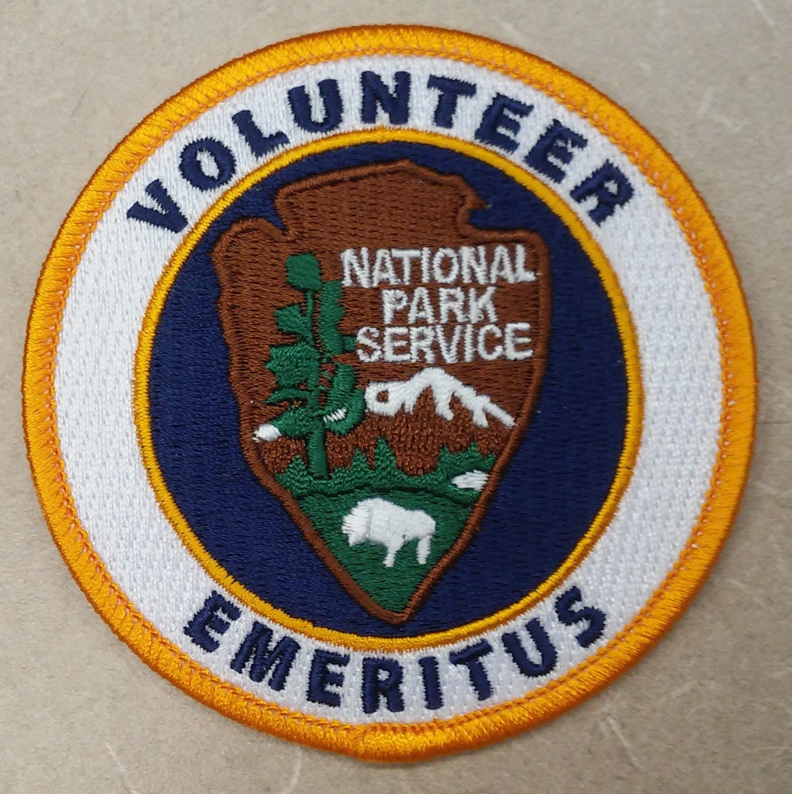 Ein kreisrunder Patch mit einem blauen Rand, weißem Hintergrund und einem gelben inneren Kreis, der zentrierte schwarzer Text trägt, der "Volunteer National Park Service Emeritus" liest.
