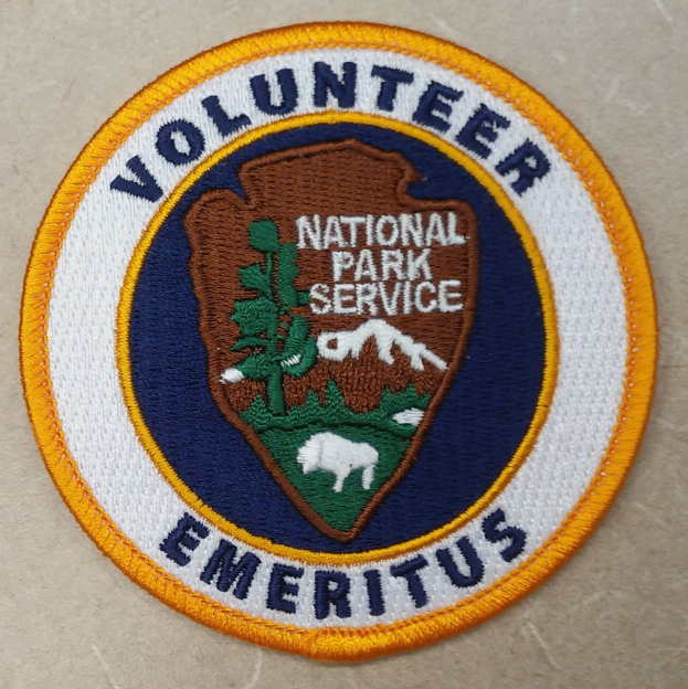 Ein kreisrunder Patch mit einem blauen Rand, weißem Hintergrund und einem gelben inneren Kreis, der zentrierte schwarzer Text trägt, der "Volunteer National Park Service Emeritus" liest.