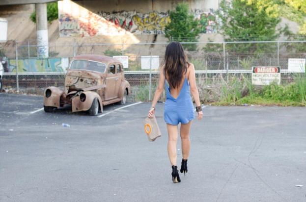 Eine Frau geht auf einer Straße und hält eine Tasche, mit einem geparkten Auto, Pflanzen, einem Gebäude und Bäumen im Hintergrund.