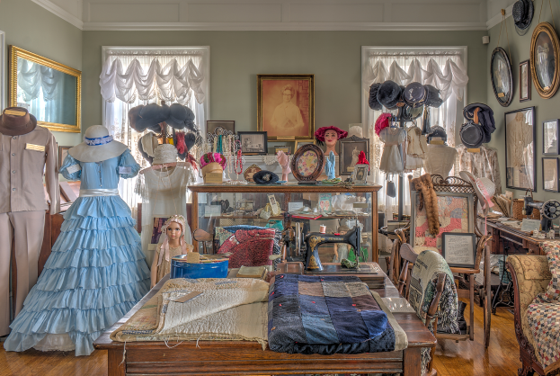 Ein Raum im Kunstmuseum von St. Louis, Missouri, voller Schaufensterpuppen in Kleidern und H├Ątchen, ein Tisch mit K├Ârpern und einer N├Ąhmaschine, St├╝hle mit Kissen, ein Schrank mit verschiedenen Gegenst├Ąnden, Fotorahmen an der Wand, Fenster mit Vorh├Ąngen und eine Decke.