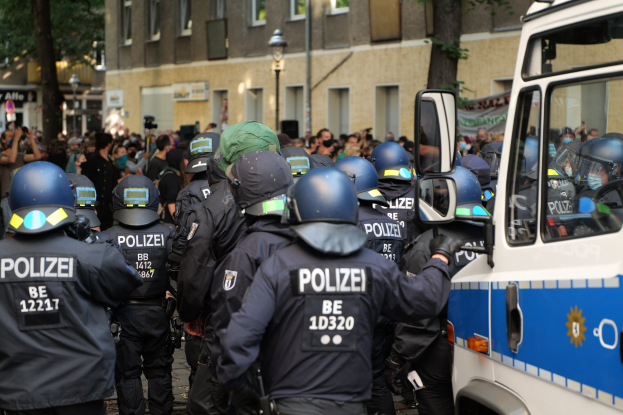 A group of police officers standing in front of a crowd, some holding guns, with a vehicle on the right and buildings, trees, and signs in the background.