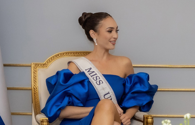 Eine Frau in einem blauen Kleid trägt einen 'Miss Universe 2019'-Schal und sitzt lächelnd auf einem Stuhl umgeben von Blumen mit einer Wand im Hintergrund.