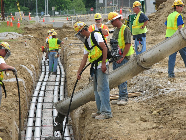 Eine Baustelle mit Arbeitern in Jacken und Helmen, Absperrbaken, Zaun, Bäume, Pfähle, Erde und Rohre im Hintergrund.