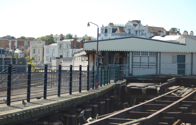 Ein Bahnhof mit einem Zug auf den Schienen, eine Brücke mit Geländern, Laternen, Verkehrssignale, Gebäude mit Fenstern, Bäume und ein Himmel im Hintergrund.