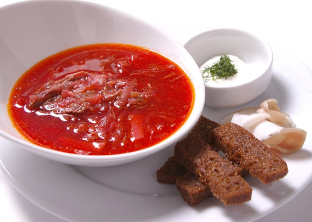 Ein weißes Tablett mit einer Schüssel dicker roter Gazpacho-Suppe, leicht getoastetes Brot und einem Zweig Petersilie als Garnitur.