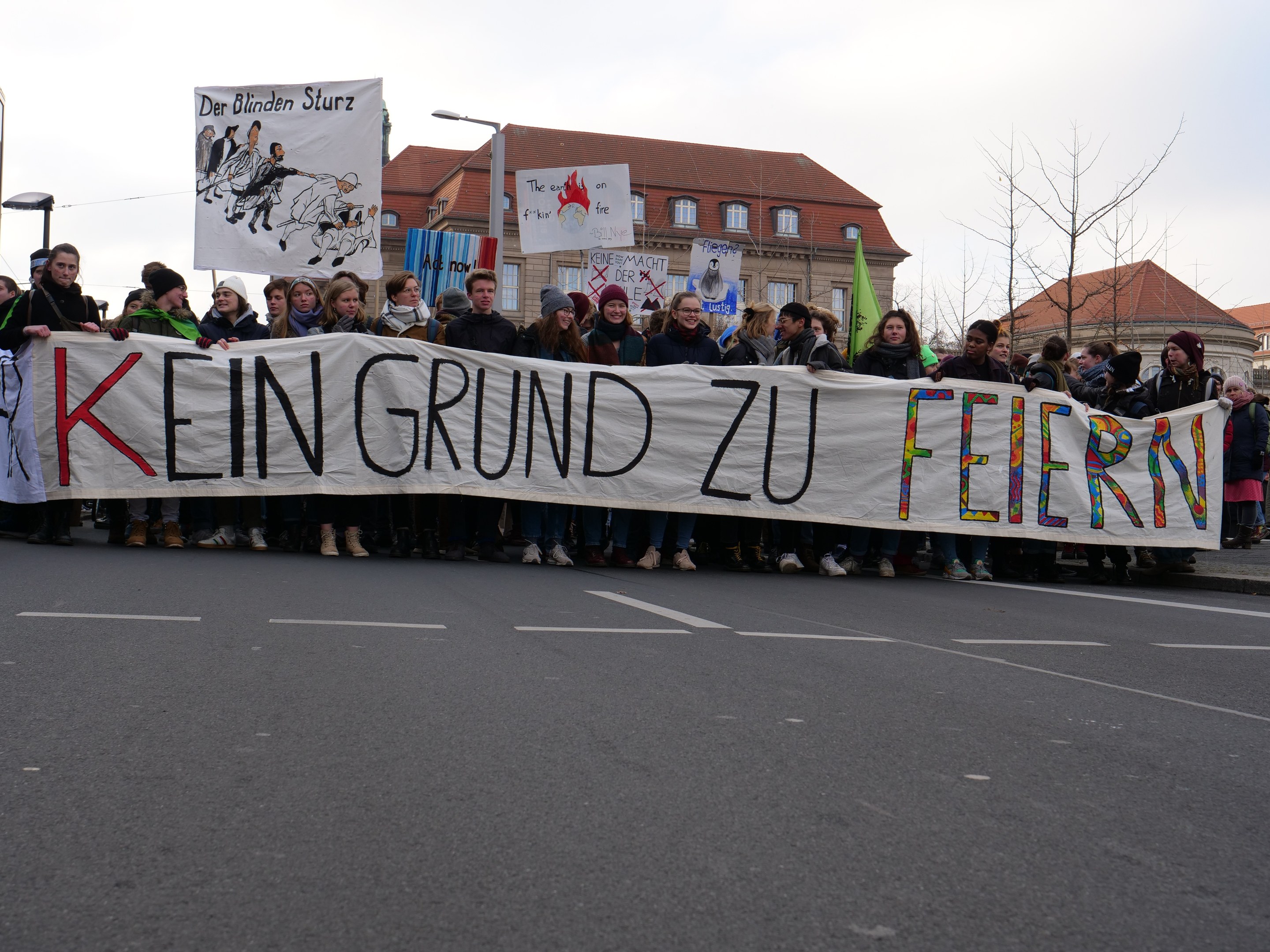 Demonstranten mit einem Transparent mit der Aufschrift 'Kein Grund zur Feier' gegen deutsche Sparmaßnahmen, im Hintergrund Straßeninfrastruktur und Gebäude.