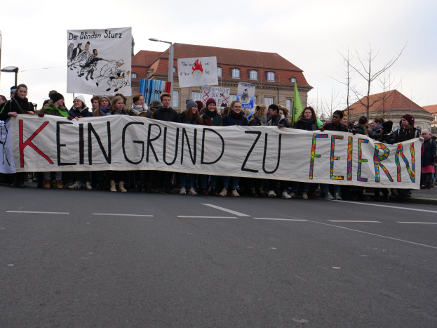 Demonstranten mit einem Transparent mit der Aufschrift 'Kein Grund zur Feier' gegen deutsche Sparmaßnahmen, im Hintergrund Straßeninfrastruktur und Gebäude.