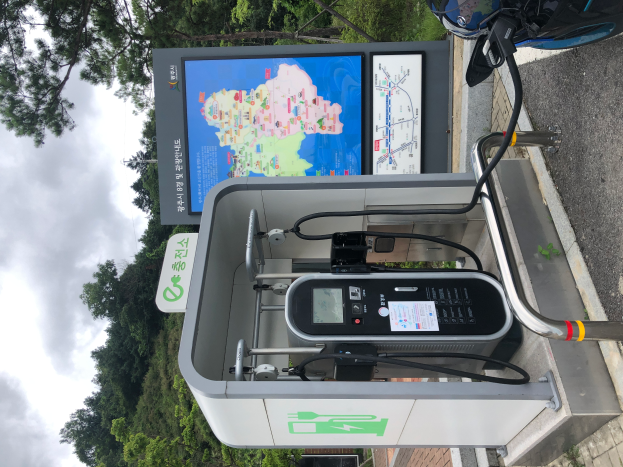Eine Ladesäule für Elektrofahrzeuge am Straßenrand mit einem Motorrad in der Nähe, Bäumen und einem klaren blauen Himmel im Hintergrund und einer digitalen Anzeigetafel, die darauf hinweist, dass es sich um eine E-Bike-Ladestation handelt.