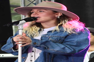 Miley Cyrus performs at the CMA Music Festival in Nashville, Tennessee, wearing a hat and singing into a microphone with a stand, with a person in a cap to her right and various musical instruments in the background.
