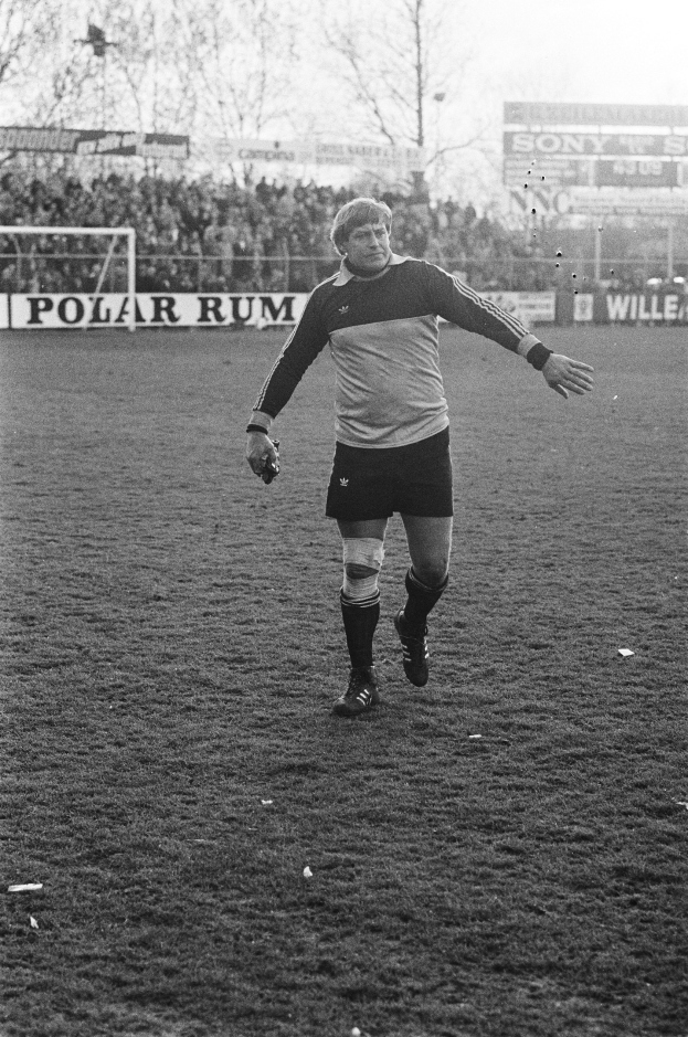 Professional soccer player in uniform standing on a field with spectators, a goal post, and trees in the background.