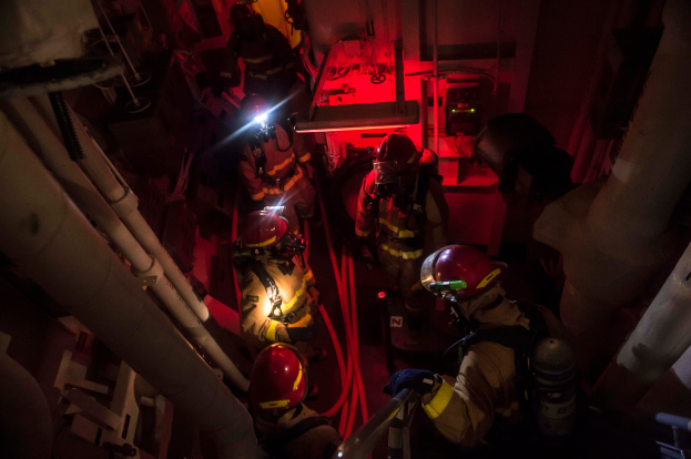 Eine Gruppe von Feuerwehrmännern in Helmen und Sauerstoffzylindern steht in einem schwach beleuchteten Raum mit Rohren auf beiden Seiten, mit einer Maschine und anderen Gegenständen im Hintergrund.