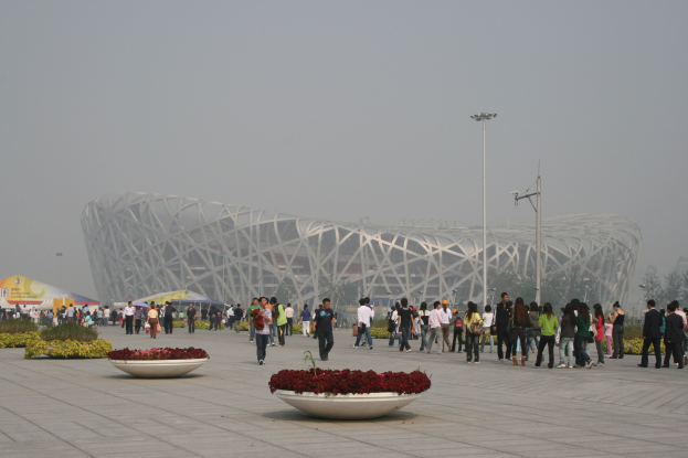 Olympiastadion in Peking, China mit Menschen, die herumlaufen, Pflanzen mit Blumen im Vordergrund, Laternenmasten, Bäumen und einem klaren blauen Himmel im Hintergrund.