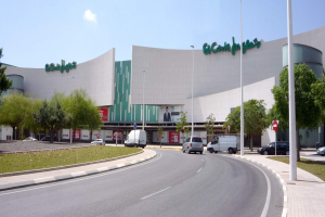 Ein großes Einkaufszentrum namens "Ecológicas" mit zahlreichen parkenden Autos davor, umgeben von Straßenlaternen, Schildern, Motorrädern, Büschen, Bäumen und einem bewölkten Himmel.