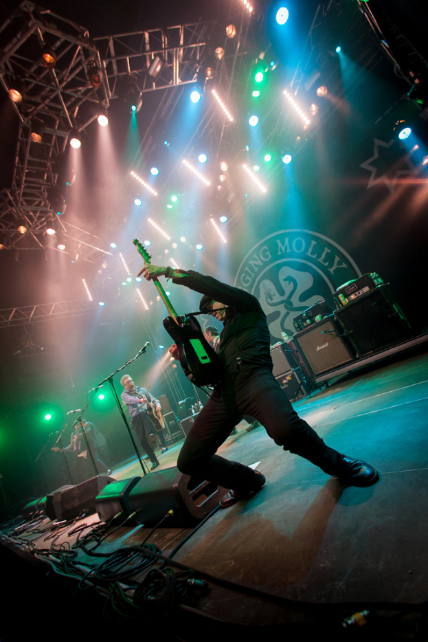 Ein musikalisches Konzert mit drei Personen auf der Bühne: zwei spielen rechts Gitarre mit Mikrofonen davor, ein Hintergrund mit "MOLLEY" und zwei Schlangenlogos, sowie Scheinwerfer über ihnen.