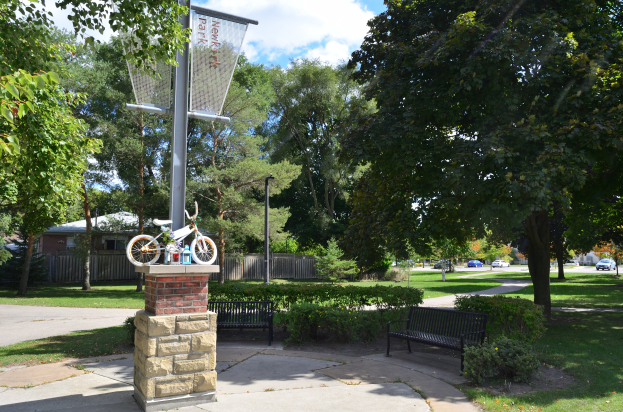 Ein Park mit einer Bank, einem Basketballkorb und einem Fahrrad an einem Pfahl, umgeben von Bäumen, Pflanzen, Gras und Häusern, mit Fahrzeugen auf einer Straße im Hintergrund unter einem bewölkten Himmel.