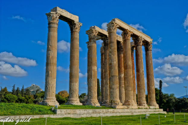 Der Tempel von Olympian Zeus in Athen, Griechenland, mit hohen Säulen, umgeben von grünem Gras, Bäumen, Menschen, Hügeln und einem bewölkten Himmel, mit Text unten.