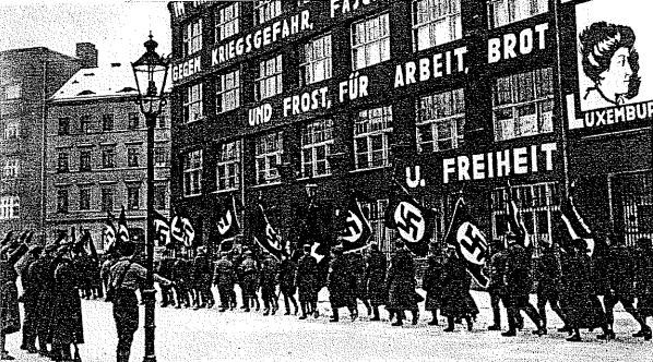 Black and white image of a group marching down a street holding flags and banners, with a light pole on the left and a Nazi Party sign in the background.