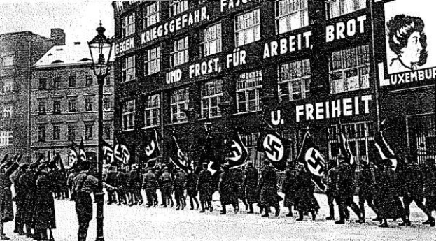 Black and white image of a group marching down a street holding flags and banners, with a light pole on the left and a Nazi Party sign in the background.