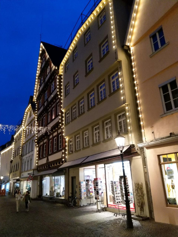 Headsteinstraße in Rothenburg ob der Tauber, Deutschland, geschmückt mit festlichen Weihnachtslichtern, mit Menschen, die spazieren gehen und einem geparkten Fahrrad, flankiert von Gebäuden mit Fenstern unter einem sichtbaren Himmel.