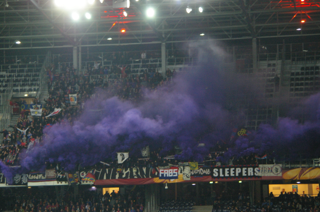 Menge in einem Stadion mit violettem Rauch, Bannern, Stühlen, Treppen, Geländern und Deckenlampen.