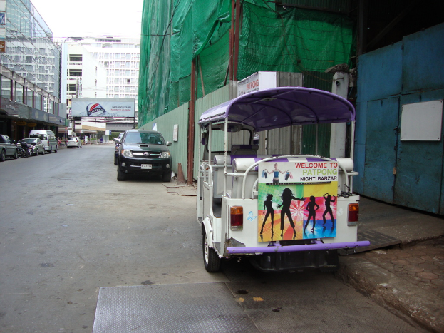 Gebäude mit parkenden Autos, eine Werbetafel, ein bewölkter Himmel und ein Auto-Rikscha am Straßenrand.