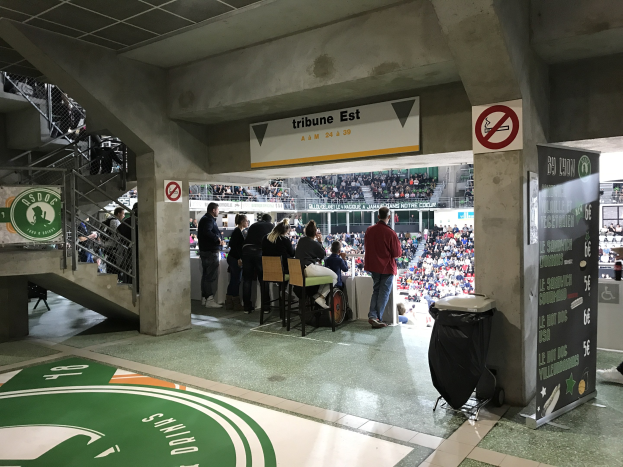 Eine Gruppe von Menschen, die vor einem sitzenden Publikum in einem Stadion mit Schildern, einem Müllcontainer, einer Texttafel, einer Treppe mit Geländern und einem Dach mit Deckenleuchten stehen; auf den Tafeln steht der Text "Boston Celtics vs New York Jets".