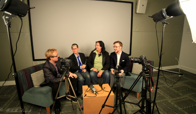 Eine Gruppe von drei Personen auf einem Sofa sitzend, wobei eine ein Mikrofon hält, gibt ein Interview vor einem Projektionsschirm, mit Kameras und Beleuchtungsausrüstung auf beiden Seiten.