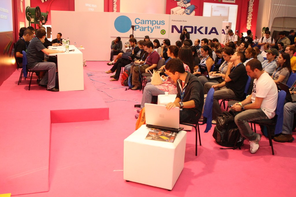 Conference with audience seated facing a stage where speakers engage in discussion, media and owners visible, with a large hoarding and wall in the background.