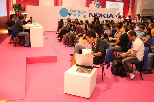 Conference with audience seated facing a stage where speakers engage in discussion, media and owners visible, with a large hoarding and wall in the background.