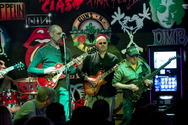 Drei Männer auf einer Bühne, die Gitarren spielen, mit einer Menge darunter und einem Graffiti oder einer Wandmalerei im Hintergrund.