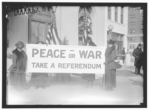 Gruppe von Frauen mit einem Banner mit der Aufschrift "Frieden oder Krieg, nehmen Sie eine Volksabstimmung" vor einem Gebäude, einer Pflanze, einem Straßenpfahl und einem bewölkten Himmel.