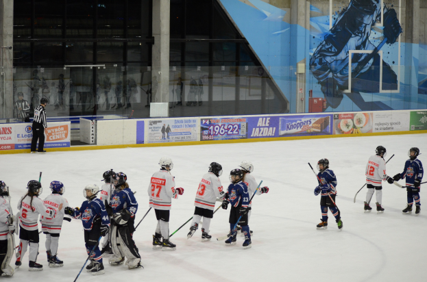 Gruppe junger Menschen in Helmen und Hockey-Ausrüstung, die auf einer Indoor-Eisfläche mit Bannern, Säulen und Glaswänden im Hintergrund Eishockey spielen.