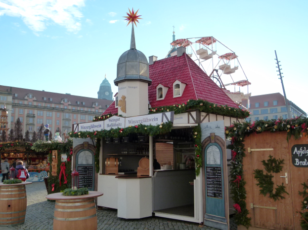 Kleines rotes Dachgebäude mit einem Riesenrad davor, umgeben von festlichen Oktoberfest-Dekorationen und Menschen, vor einer Kulisse aus Gebäuden, Bäumen und bewölktem Himmel.