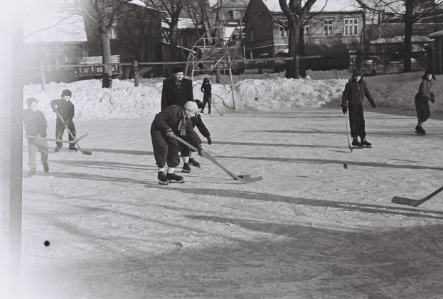 Eine Gruppe von Menschen, die Hockey auf einem Eisplatz spielen, umgeben von Schnee, Bäumen, Gebäuden mit Fenstern, Treppen mit Geländern und einem Lastwagen im Hintergrund, dargestellt in Schwarz-Weiß.