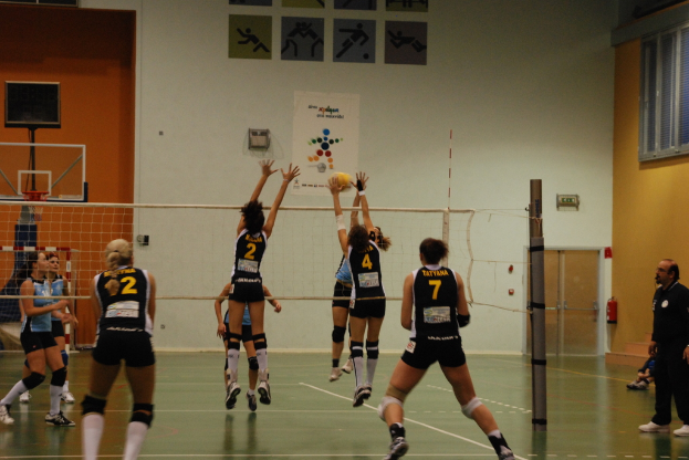 Ein Volleyballspiel auf einem Indoor-Court, mit aktiven Spielern, einem Basketballkorb im Hintergrund und einer Tür und Fenstern an den Wänden.