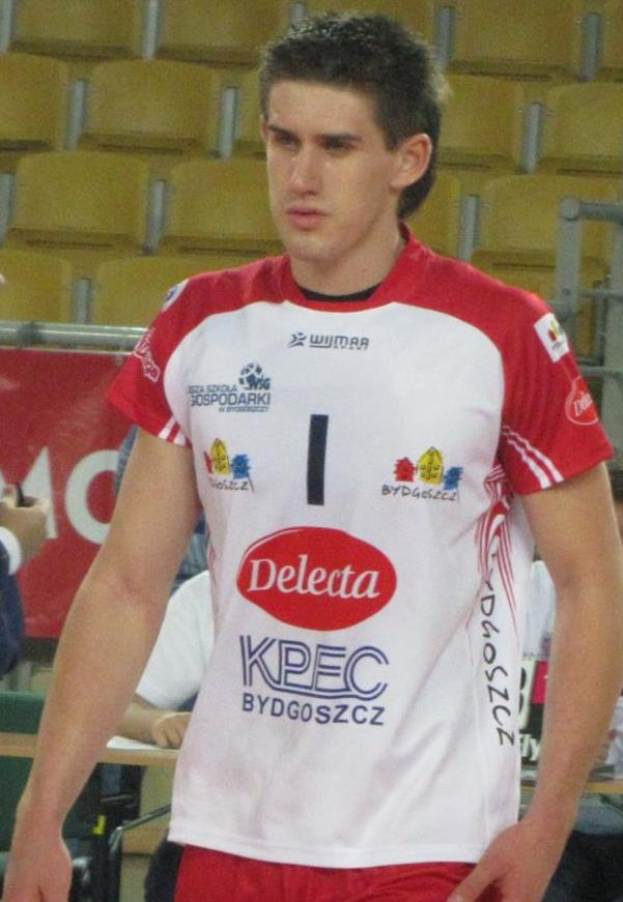 Ein Mann in einem roten und weißen Handballtrikot steht auf einem Feld, umgeben von Zuschauern, mit einem roten Banner im Hintergrund, das "Krasnoyarsk vs Bydgoszcz" zeigt und entschlossen aussieht.