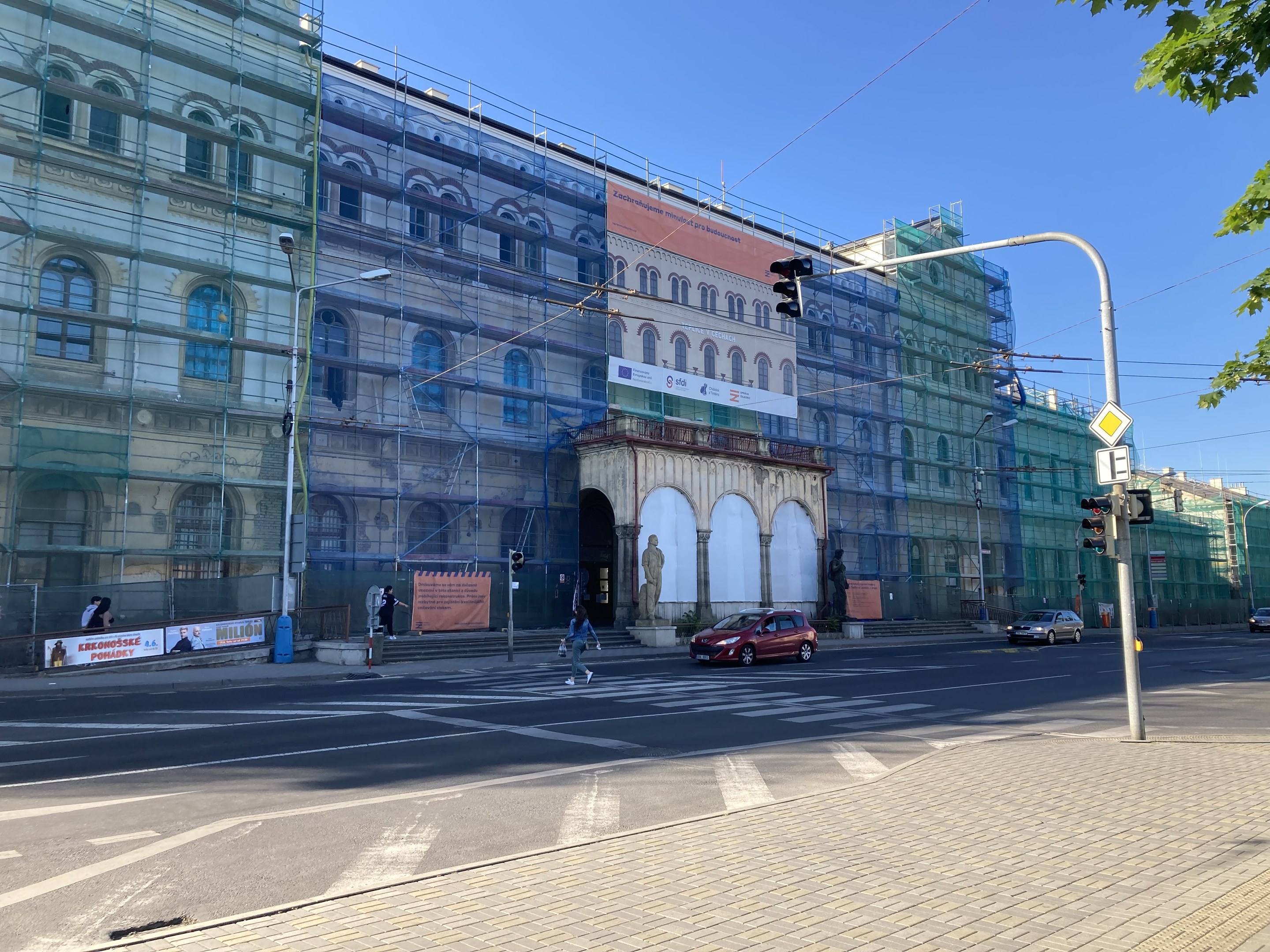 Großes Gebäude mit Gerüst, das renoviert wird, umgeben von Fahrzeugen, Fußgängern, Ampeln, Schildern, Straßeninfrastruktur, Bäumen und einem klaren blauen Himmel.
