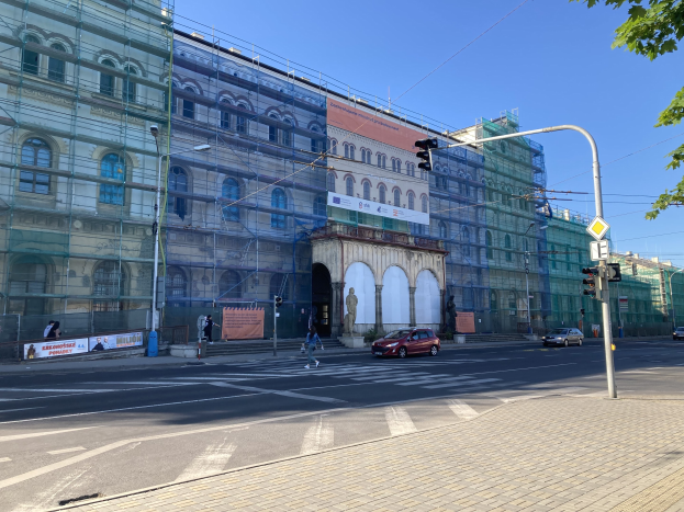Großes Gebäude mit Gerüst, das renoviert wird, umgeben von Fahrzeugen, Fußgängern, Ampeln, Schildern, Straßeninfrastruktur, Bäumen und einem klaren blauen Himmel.