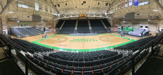 Eine geräumige Turnhalle mit einem zentralen Basketballfeld, umgeben von Stühlen, Geländern, Treppen, Bannern, Bildschirmen, Lampen und anderen Gegenständen, mit dem Boden unten sichtbar.