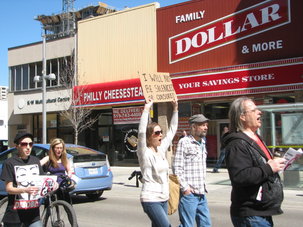 Eine Gruppe von Menschen, die auf einer StraÃe protestieren, mit GebÃ¤uden, einem Mast und einem Baum im Hintergrund, einem Auto und einem Fahrrad auf der linken Seite und dem Himmel oben sichtbar.