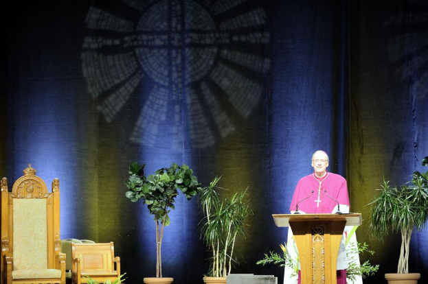 Ein Mann in einem rosaweißen Kleid steht neben einem Rednerpult in einem Auditorium, mit Pflanzen und zwei Stühlen daneben und einer bunten Wand im Hintergrund.