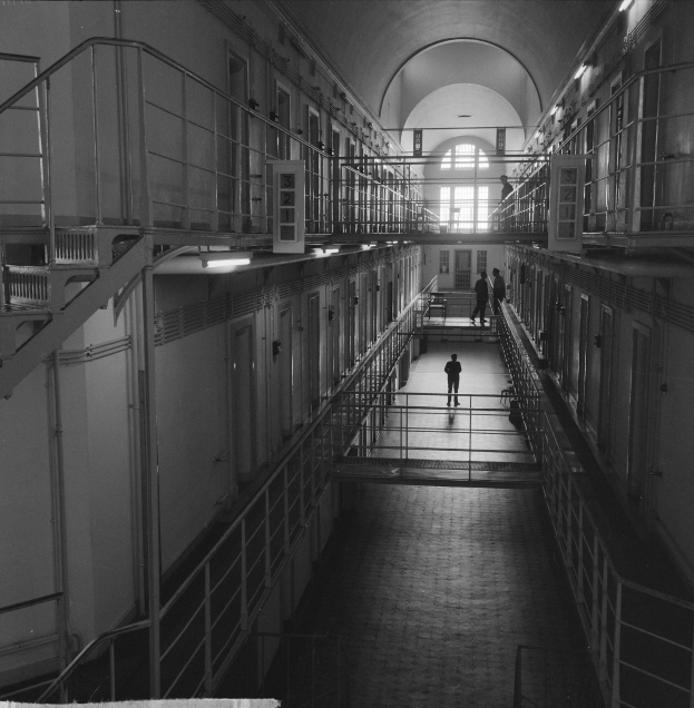 Schwarzes und weißes Foto eines Mannes, der einen langen Gefängnishofgang mit Gittern, Türen, Lampen und einem Fenster im Hintergrund entlanggeht.