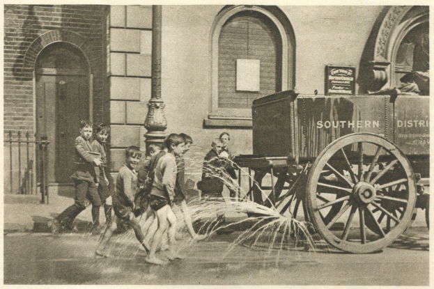 Schwarz-weiß-Foto von Kindern, die mit einem Feuerhydranten vor einem Gebäude mit Fenstern, einer Tür, einem Mast und einem Zaun spielen, mit einem Wagen im Hintergrund.