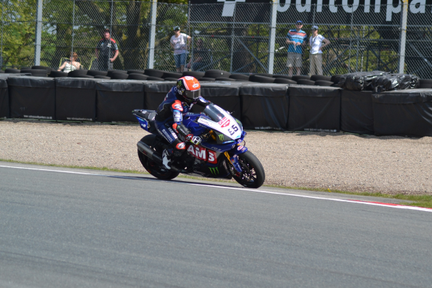 Ein Motorradfahrer in Helm und Handschuhen fährt auf einer von Gras, Reifen, Maschendrahtzaun und einem Banner umgebenen Rennstrecke, mit Bäumen und einem klaren blauen Himmel im Hintergrund.